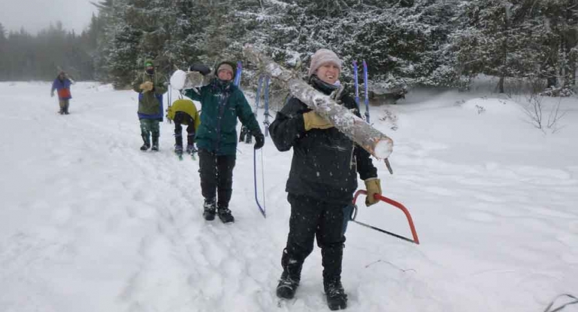 a group of gap year students carry a felled tree and saws across a snowy landscape